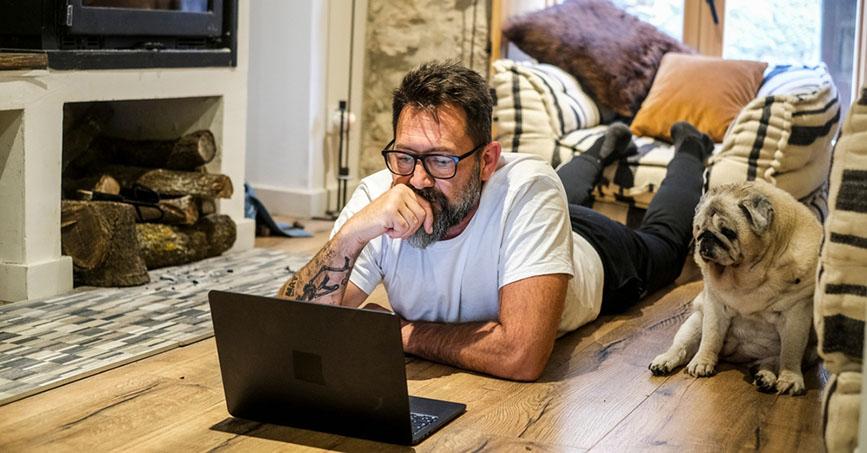 A man laying on the floor looking at his laptop with a dog next to him. 