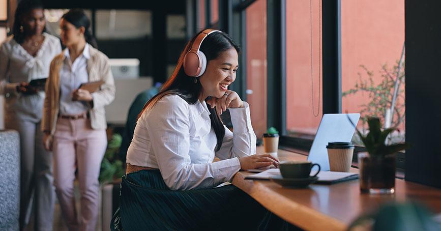 Smiling woman wearing headphones working on a laptop at a window-side counter in a cafe, with coffee and a plant beside her; two women walking and talking in the background.