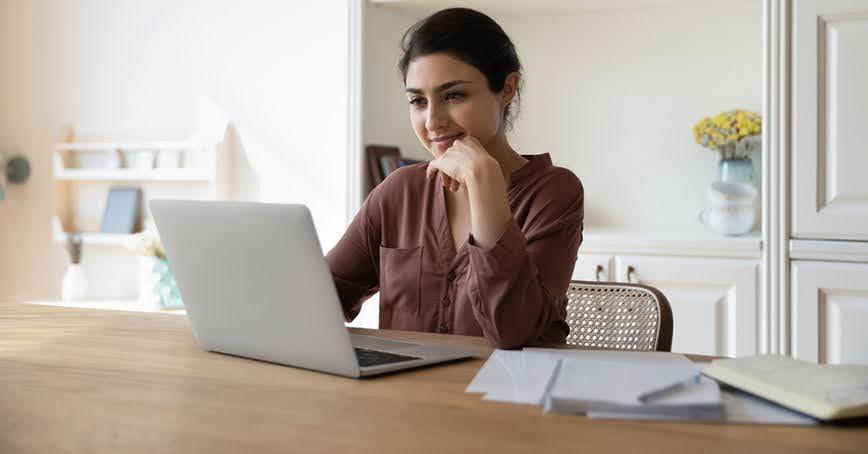 A woman with dark hair, wearing a brown shirt, smiles while looking at her laptop at a kitchen table.