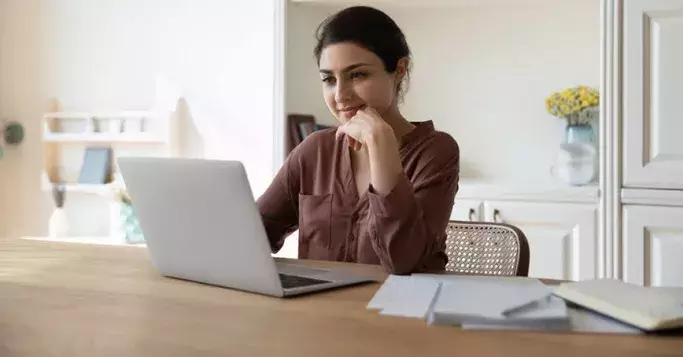 A woman with dark hair, wearing a brown shirt, smiles while looking at her laptop at a kitchen table.