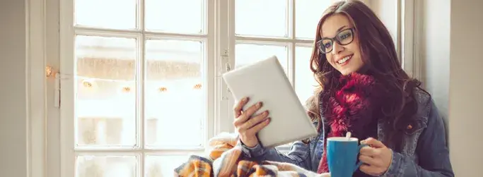 Woman enjoying streaming on a tablet while relaxing with a warm drink by the window.